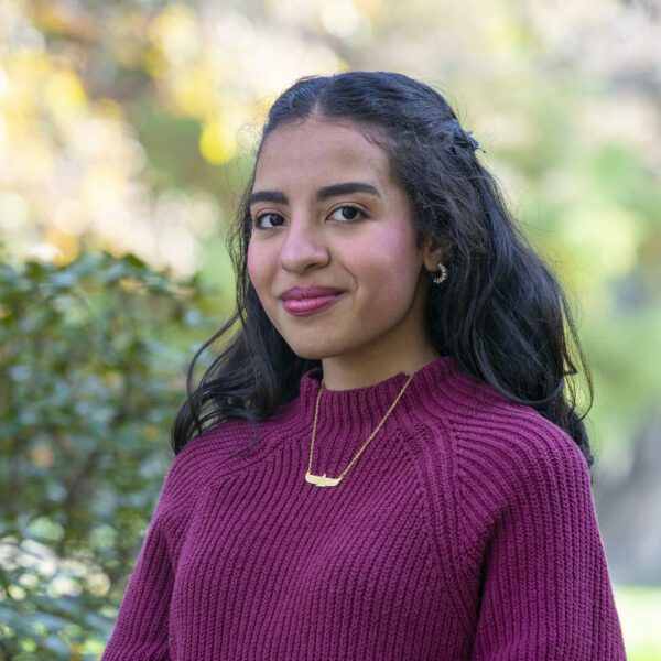 Nayera Hasan ’26 stands outdoors on campus in a burgundy sweater and gold necklace, with trees and greenery softly blurred in the background.