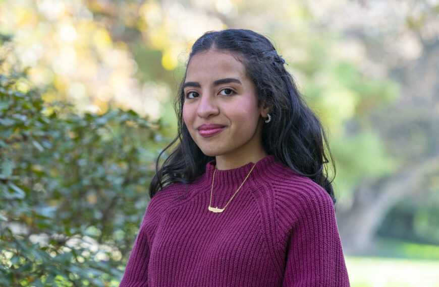 Nayera Hasan ’26 stands outdoors on campus in a burgundy sweater and gold necklace, with trees and greenery softly blurred in the background.