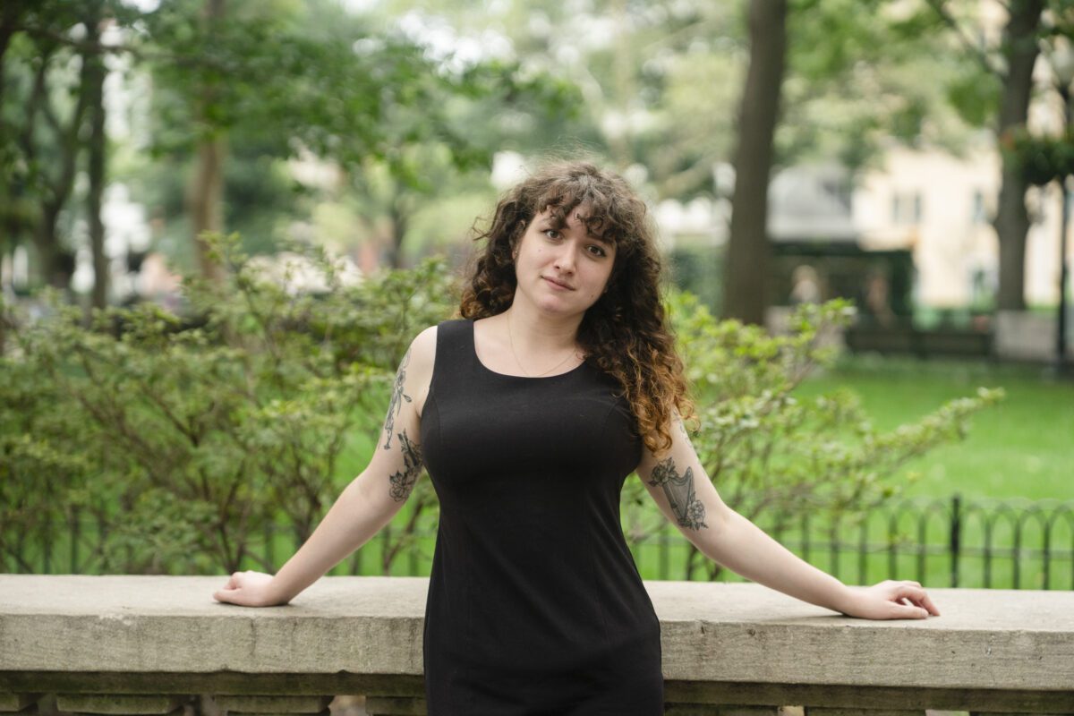 Sarah Weill-Jones ’26 stands outdoors on campus in a black sleeveless dress, leaning against a stone railing with trees and shrubs in the background.