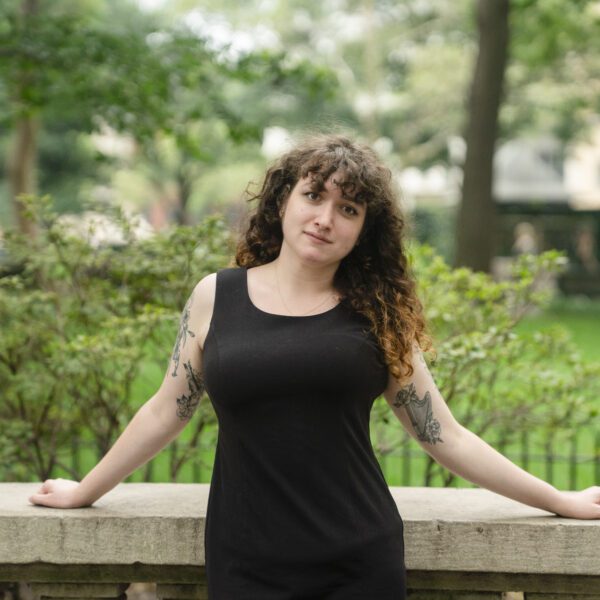 Sarah Weill-Jones ’26 stands outdoors on campus in a black sleeveless dress, leaning against a stone railing with trees and shrubs in the background.