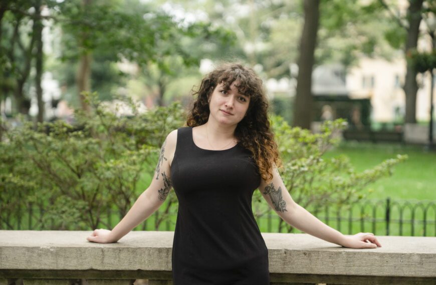Sarah Weill-Jones ’26 stands outdoors on campus in a black sleeveless dress, leaning against a stone railing with trees and shrubs in the background.