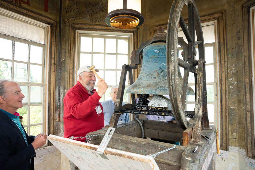 An alum raises a mallet to ring an old bell inside a weathered bell tower covered with signatures and messages.