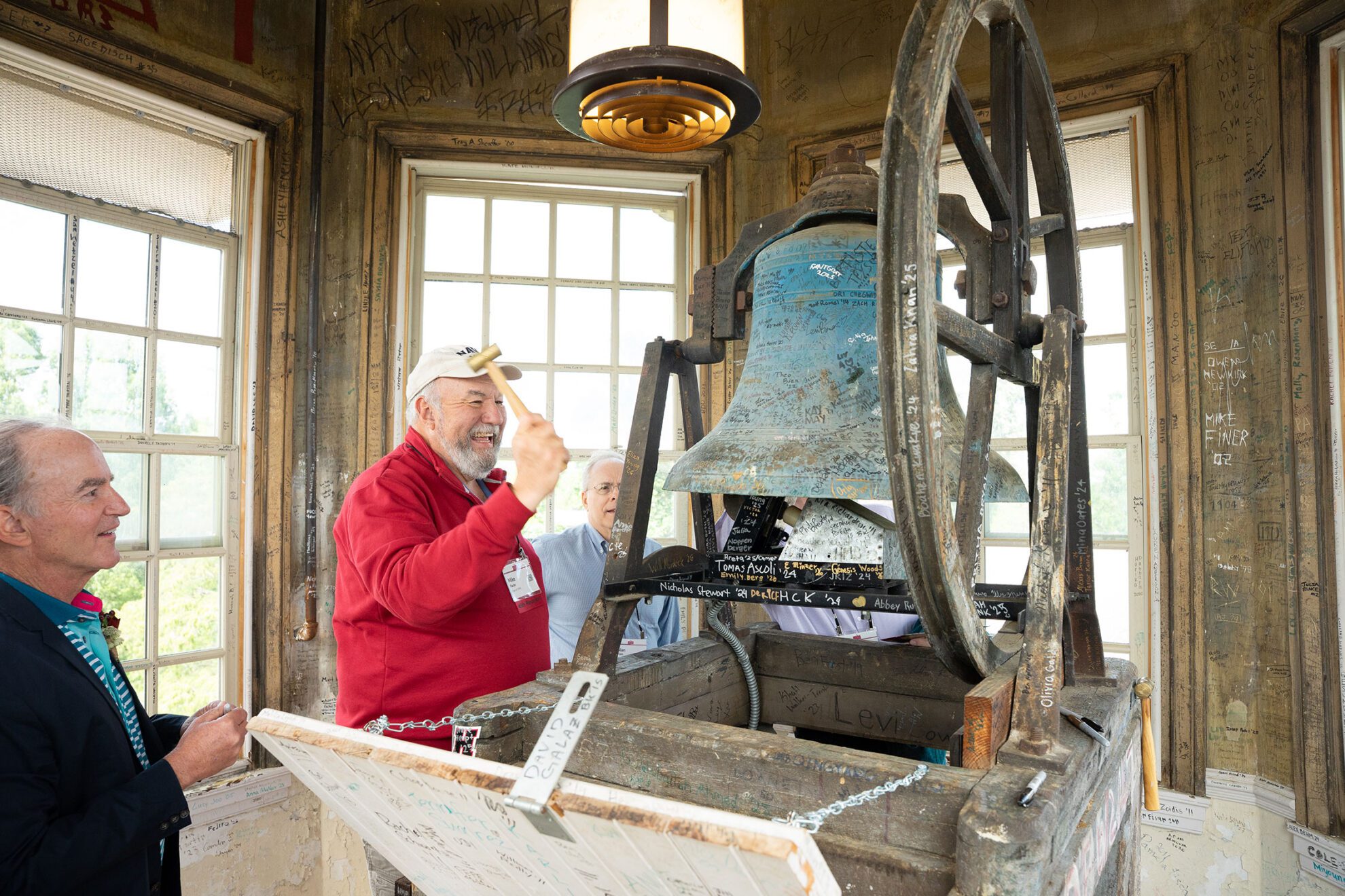An alum raises a mallet to ring an old bell inside a weathered bell tower covered with signatures and messages.