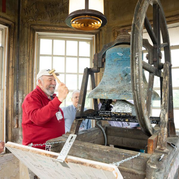 An alum raises a mallet to ring an old bell inside a weathered bell tower covered with signatures and messages.
