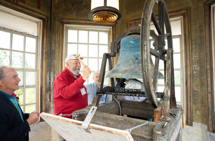 An alum raises a mallet to ring an old bell inside a weathered bell tower covered with signatures and messages.