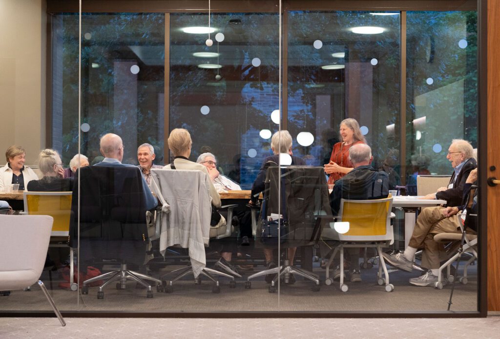 Reunion attendees sit around a large table in a glass-walled meeting room, listening as one woman stands and speaks to the group.