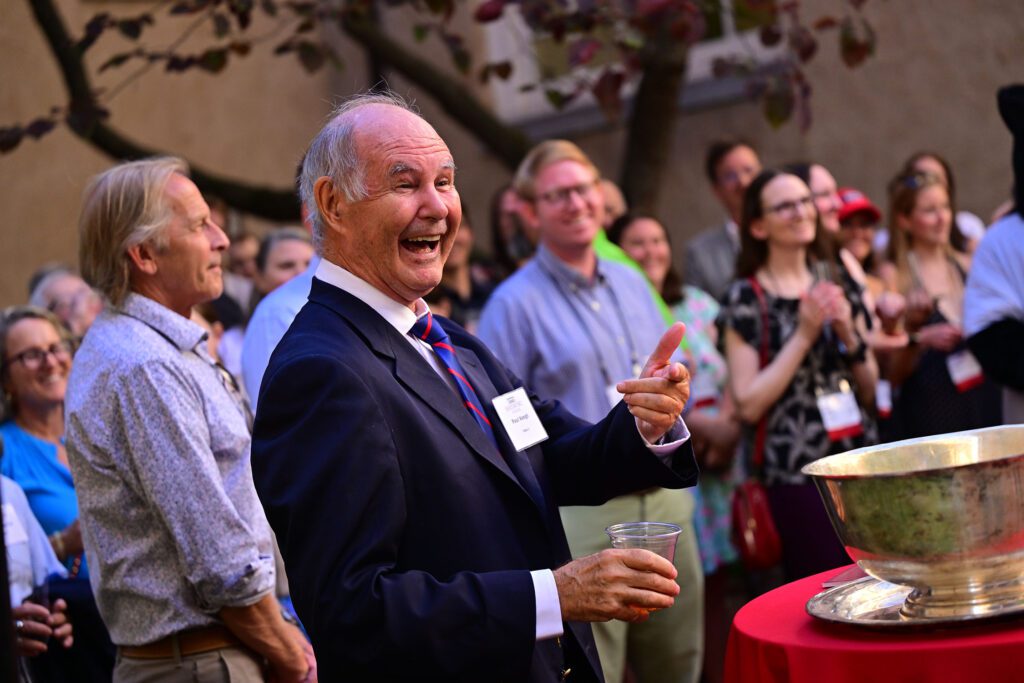 An alum in a suit laughs and gestures with a drink in hand while a crowd of attendees smiles behind him at an outdoor reception.