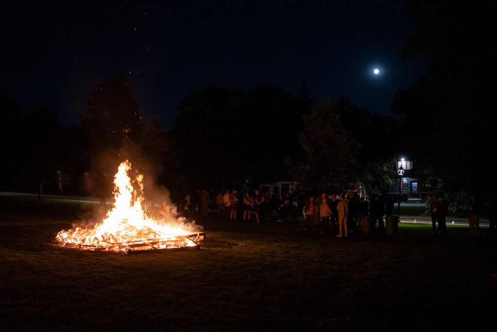A large bonfire burns on the lawn at night as people gather nearby under a bright moon.