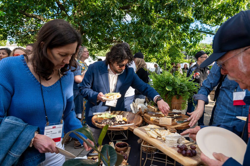 Alumni fill plates from a buffet table outdoors beneath leafy trees during a cocktail reception.