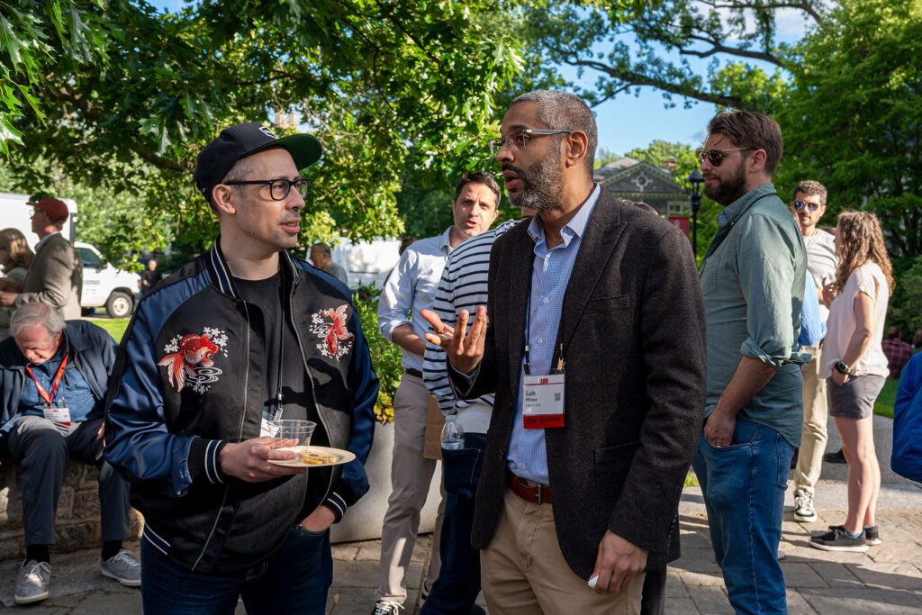 Two alumni stand talking closely beneath trees during an outdoor reception while other guests mingle nearby.