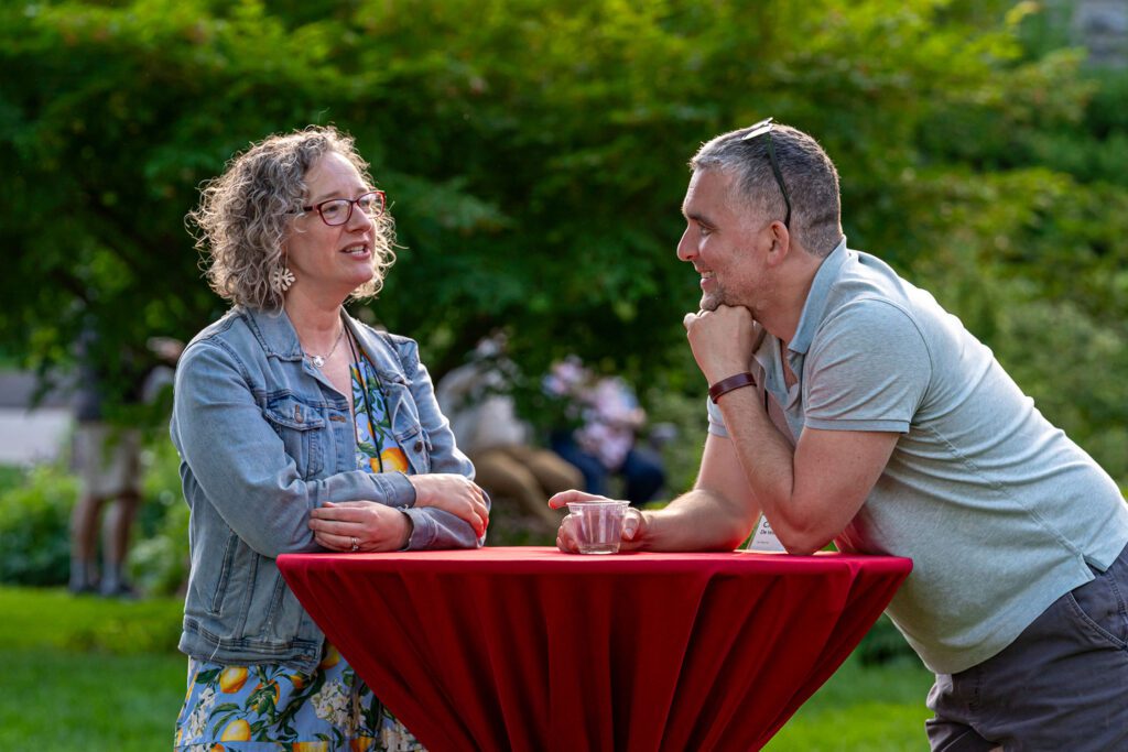 Two alumni lean against a tall red cocktail table while talking in a garden setting at dusk.