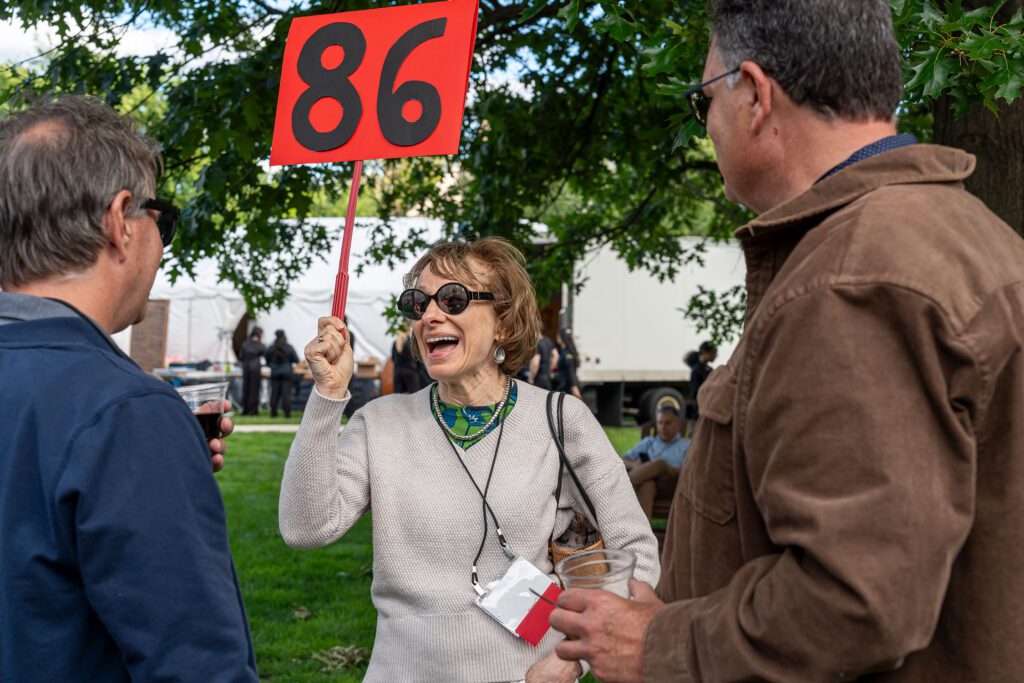 A smiling alum wearing sunglasses holds up a red sign with the number 86 while talking with two other attendees.