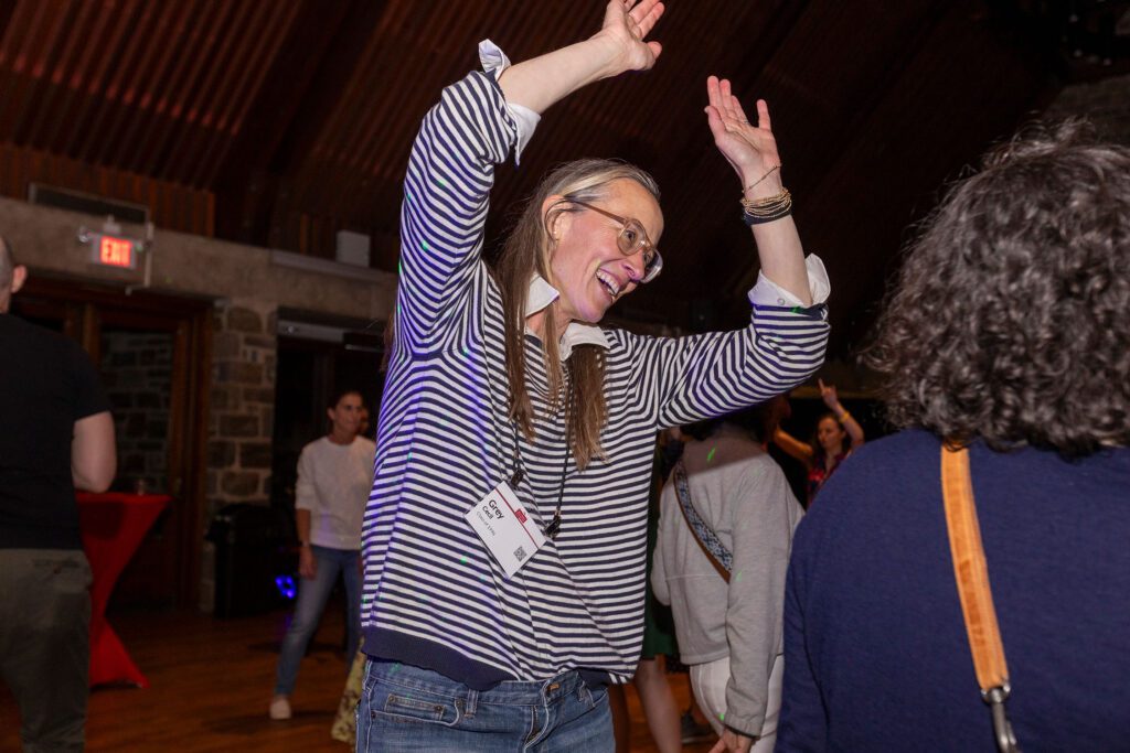 An alum in a striped shirt dances with both arms raised inside a wood-paneled room as others mingle nearby.