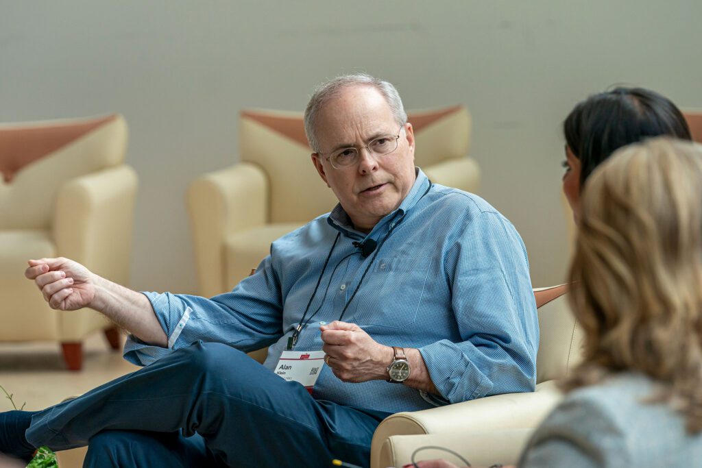 A man seated in an armchair gestures while speaking during a panel discussion, with two audience members visible in the foreground.
