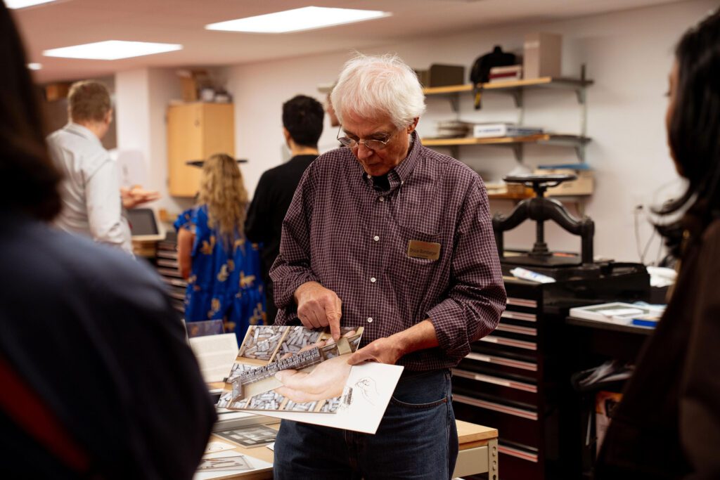 A man stands in a lab-like workspace holding and pointing to a printed sheet as visitors gather around to listen.
