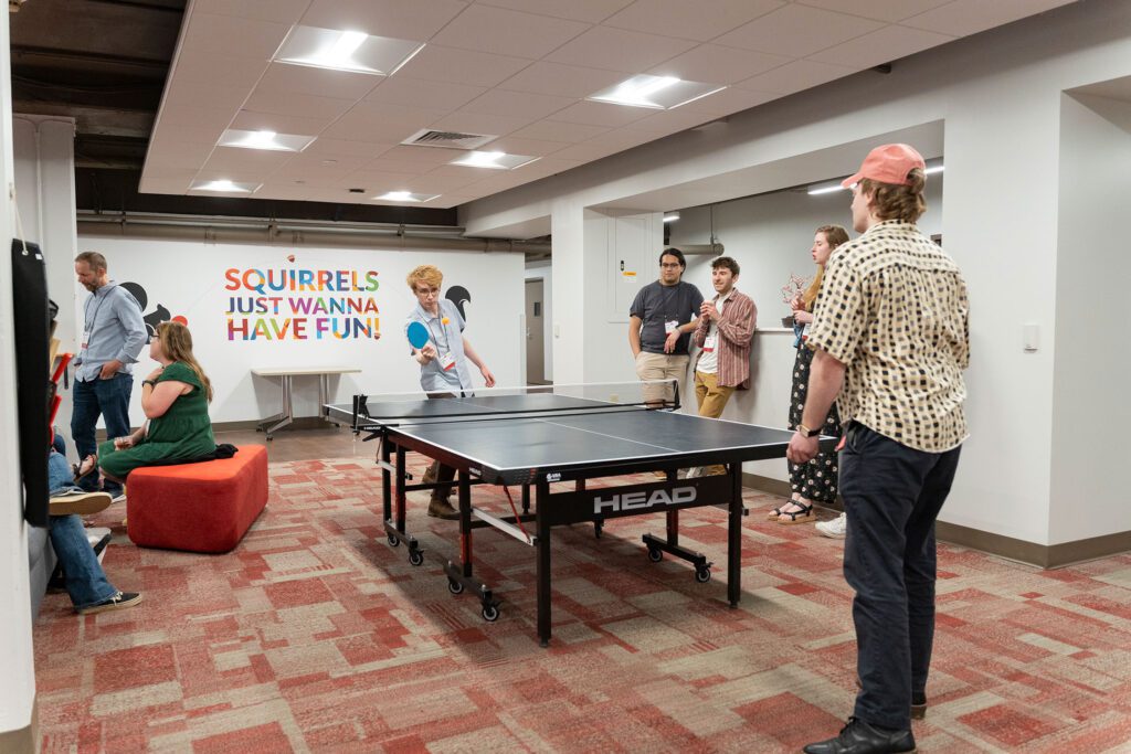 Alumni and guests gather around a ping-pong table in a lounge as two people play a game and others watch from the sidelines beneath a colorful wall sign reading “Squirrels Just Wanna Have Fun!”