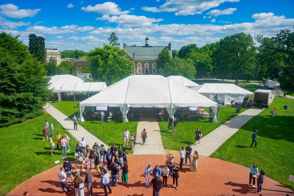 A wide overhead view shows large white event tents on the lawn as alumni and guests gather on brick walkways and grass beneath a bright blue sky with scattered clouds.