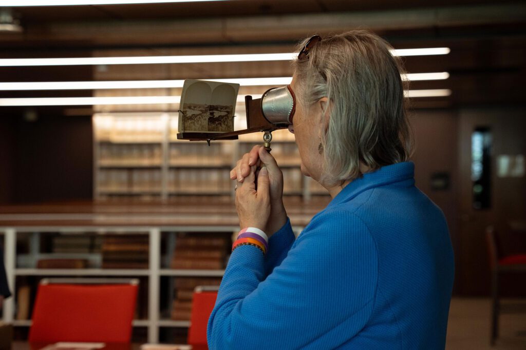 An alum in a blue top looks through a handheld stereoscope viewer at an old stereographic image inside a library or archive space.