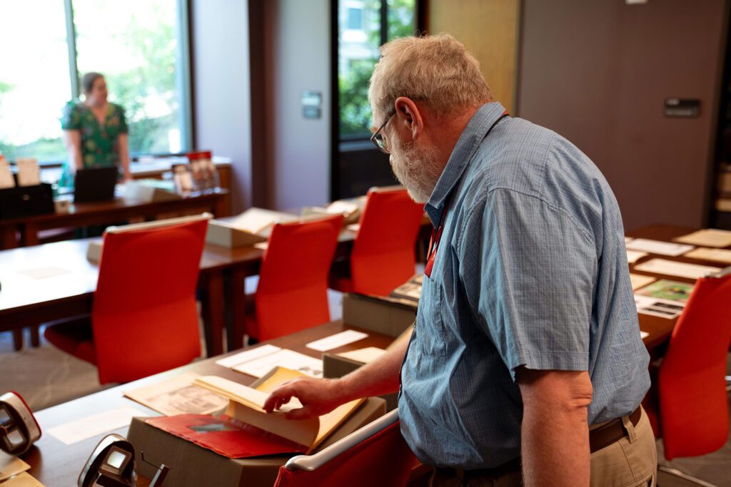 An attendee leans over a table examining an open archival volume in a room set with red chairs and display materials, while another person stands in the background.