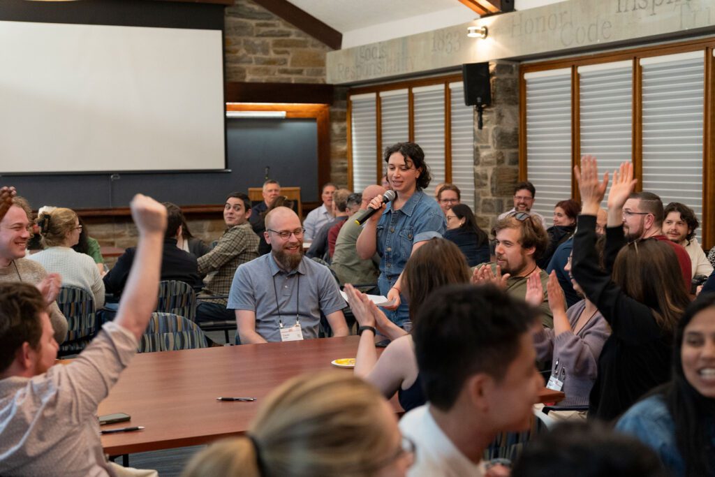 A person holding a microphone stands among tables of reunion attendees as many participants raise their hands during a lively trivia event.