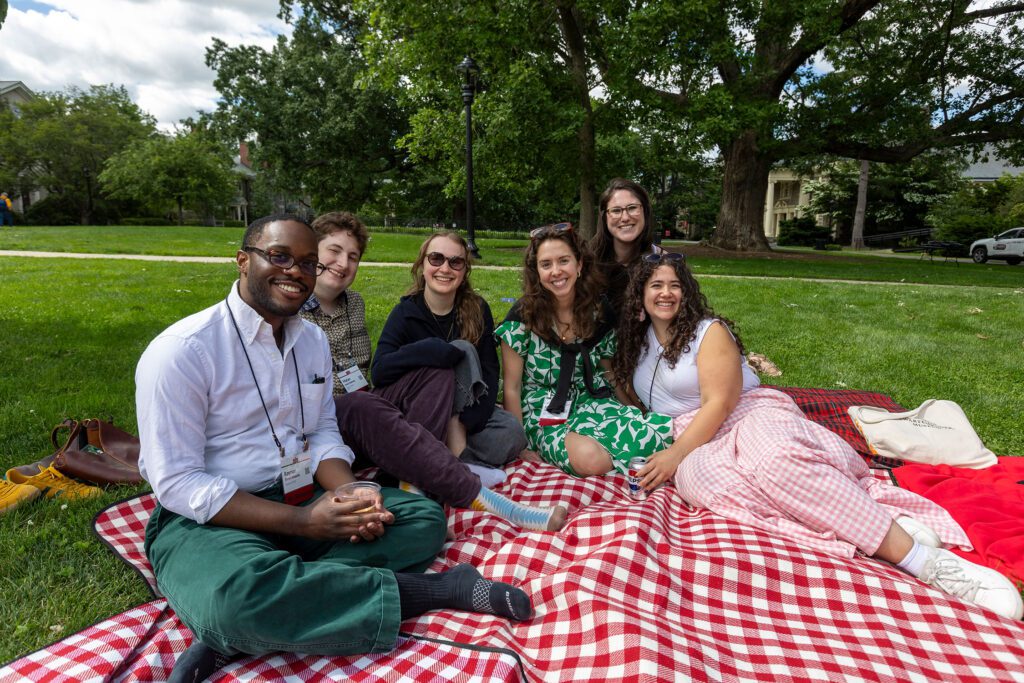 Six alumni sit together on a red-and-white picnic blanket on the lawn, smiling at the camera with trees and campus buildings behind them.