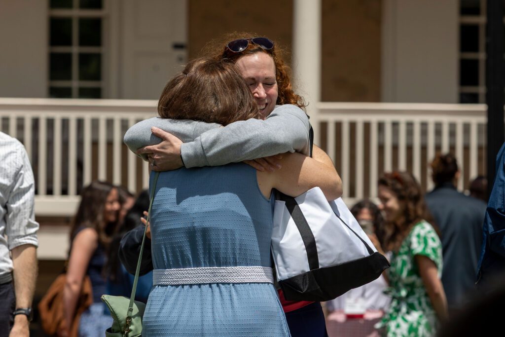 Two people embrace warmly outdoors in front of a campus building while other reunion guests mingle in the background.