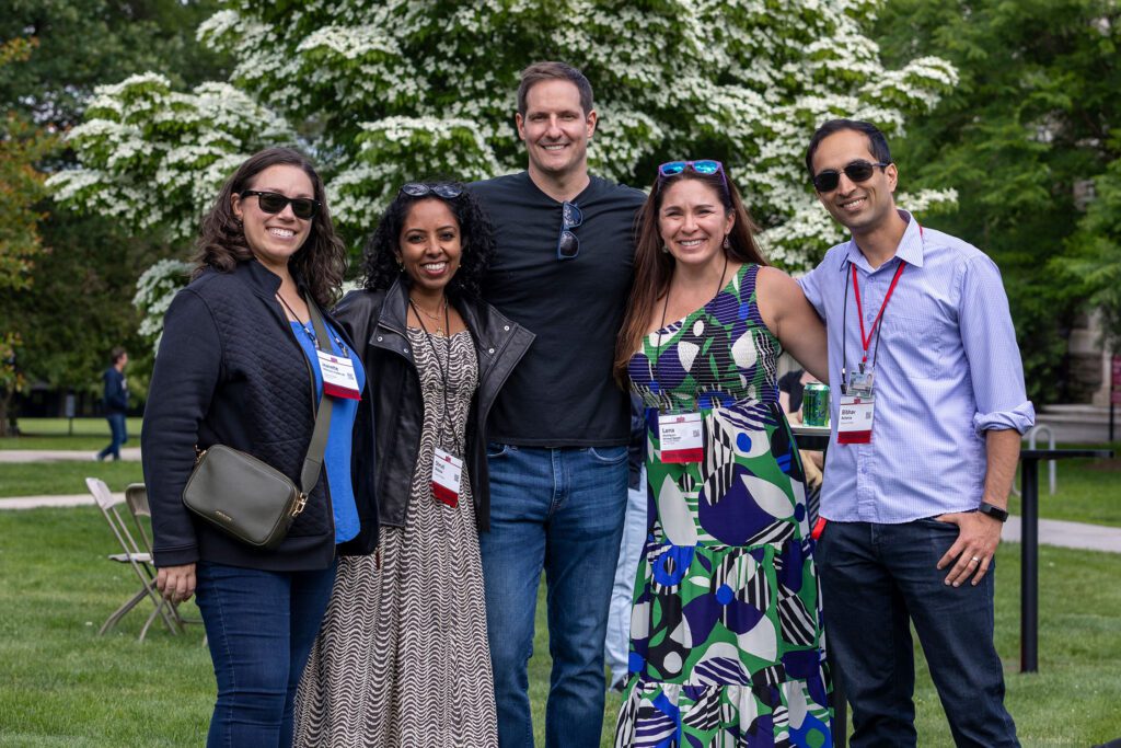 Five alumni stand arm in arm smiling for a group photo on the lawn in front of a flowering tree.