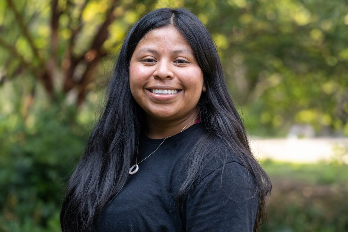 Izzy Muraca ’26 smiles outdoors on campus, wearing a black top and silver pendant necklace against a softly blurred background of trees and greenery.