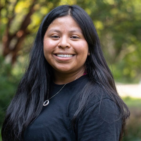 Izzy Muraca ’26 smiles outdoors on campus, wearing a black top and silver pendant necklace against a softly blurred background of trees and greenery.