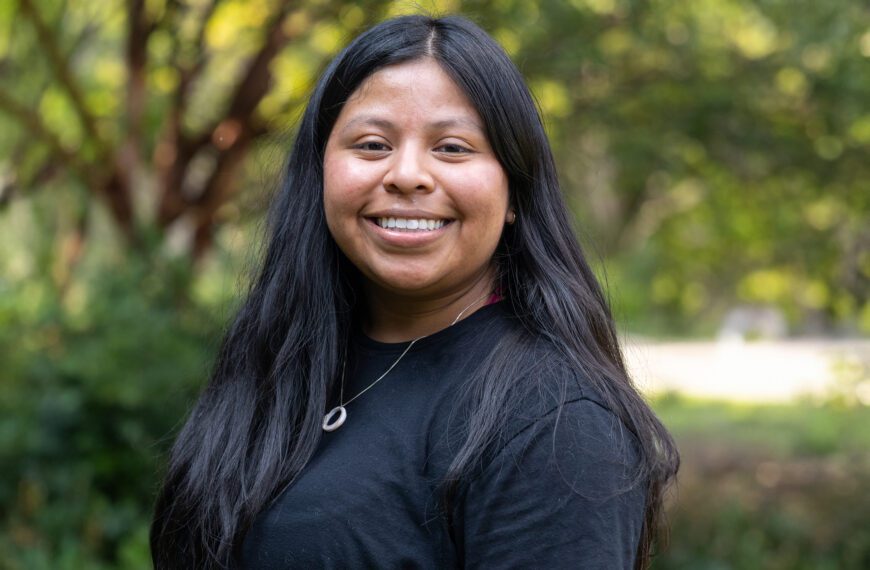 Izzy Muraca ’26 smiles outdoors on campus, wearing a black top and silver pendant necklace against a softly blurred background of trees and greenery.
