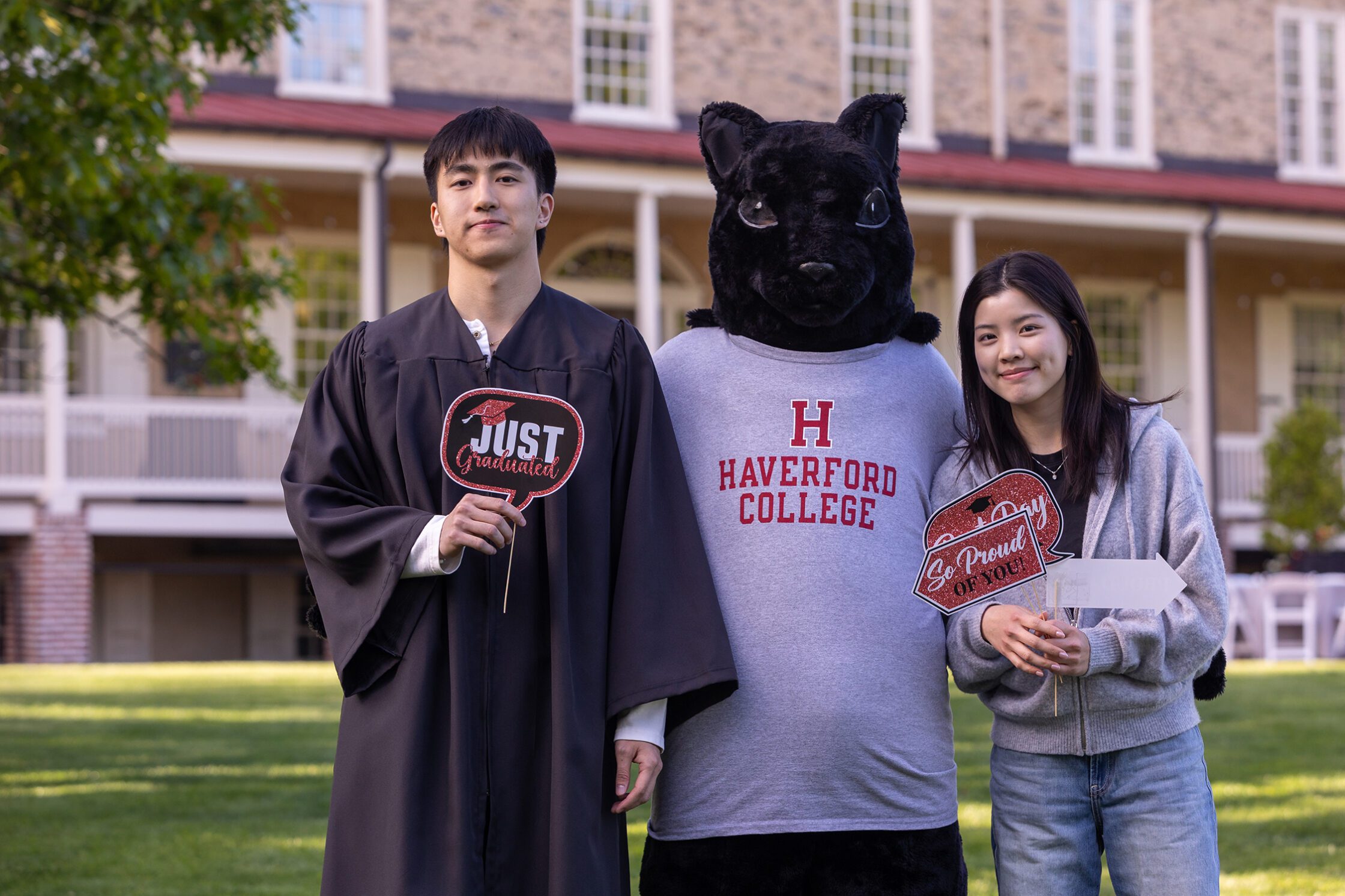 Daniel Lee stands on the lawn with the Haverford squirrel mascot and a smiling supporter, holding celebratory graduation signs in front of Founders Hall.