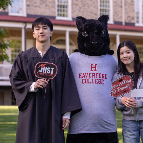 Daniel Lee stands on the lawn with the Haverford squirrel mascot and a smiling supporter, holding celebratory graduation signs in front of Founders Hall.