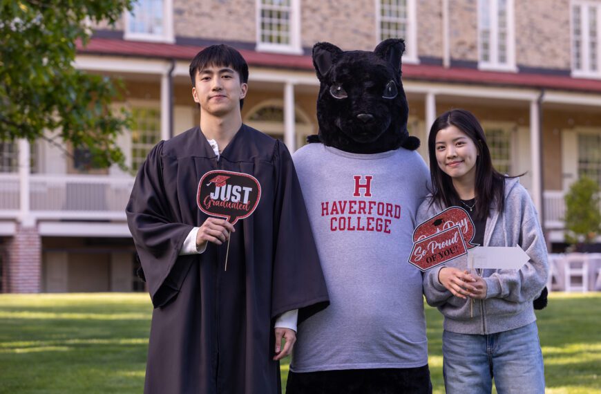 Daniel Lee stands on the lawn with the Haverford squirrel mascot and a smiling supporter, holding celebratory graduation signs in front of Founders Hall.