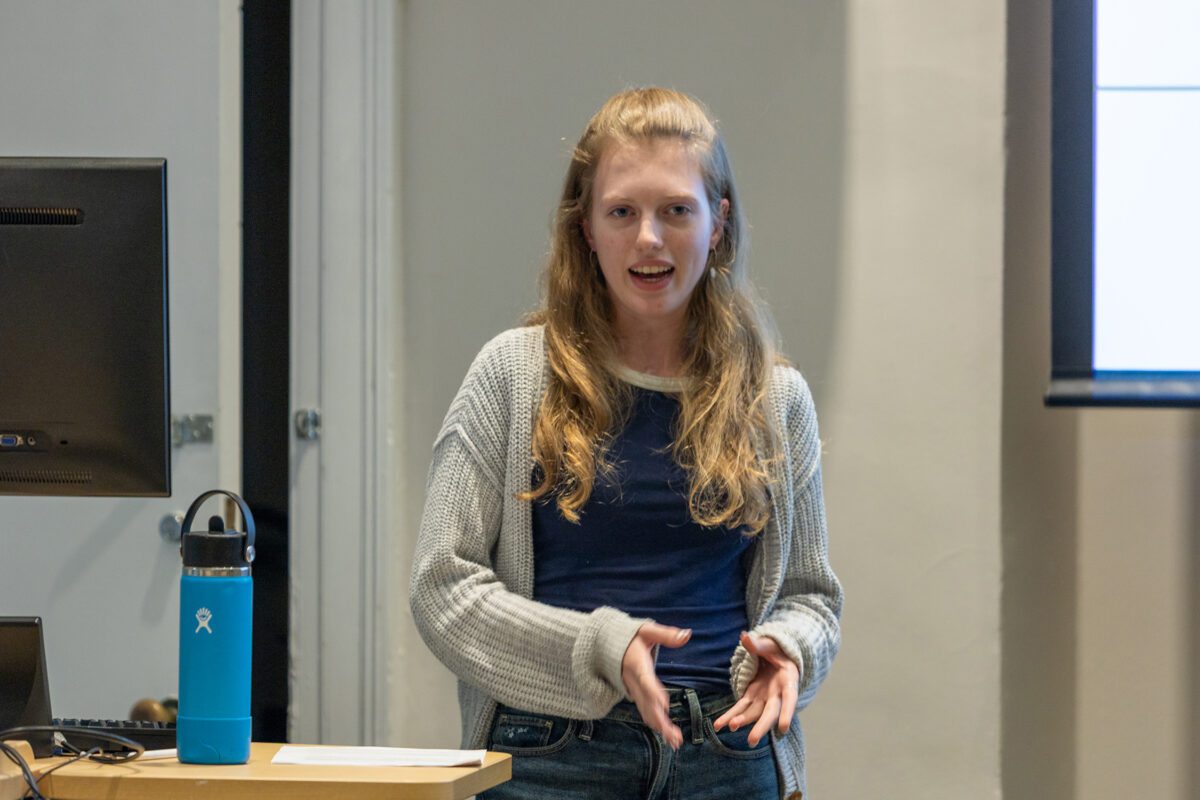 A student stands at the front of a classroom giving a presentation, speaking with her hands near a lectern and a blue water bottle beside a computer monitor.