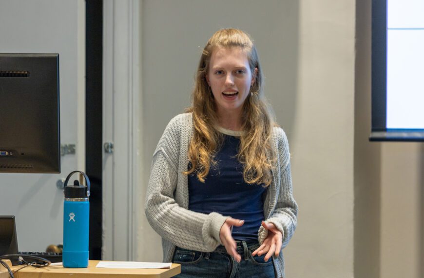 A student stands at the front of a classroom giving a presentation, speaking with her hands near a lectern and a blue water bottle beside a computer monitor.